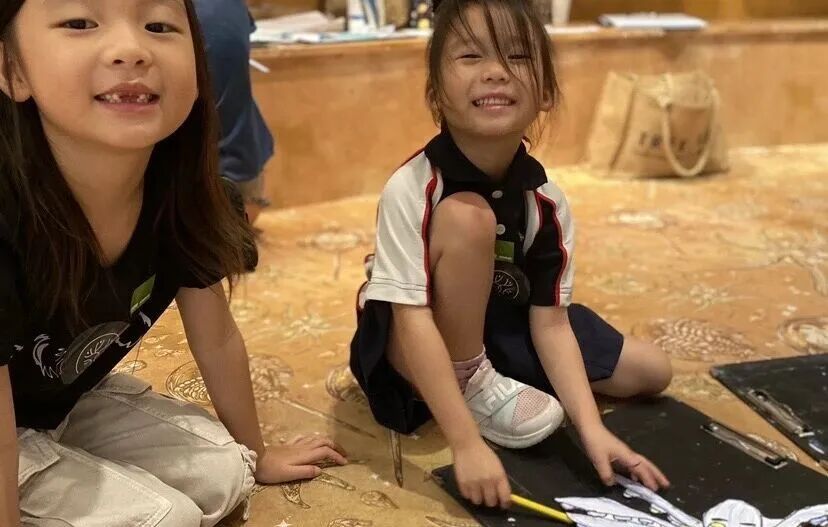 Two girls sit on a patterned carpet drawing on clipboards