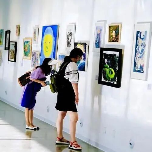 Two visitors lean in to look closely at framed artworks along a bright gallery wall