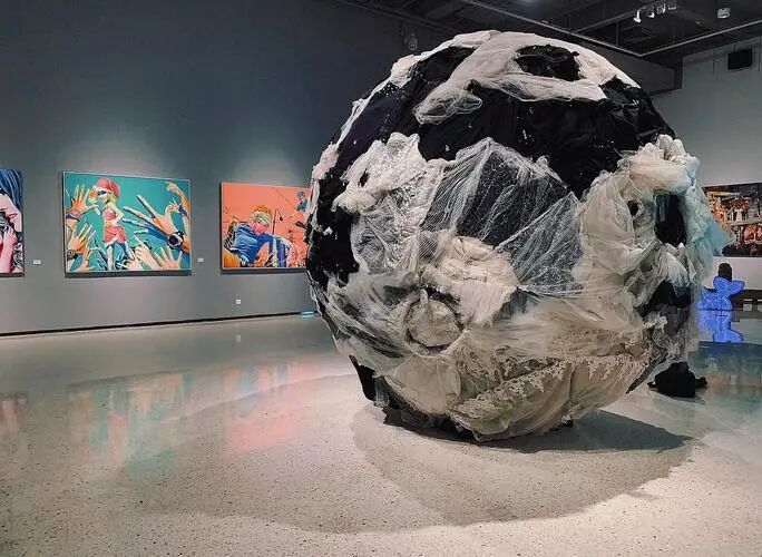A large sphere sculpture wrapped in black and white fabric stands in a gallery lined with colourful pop-art paintings