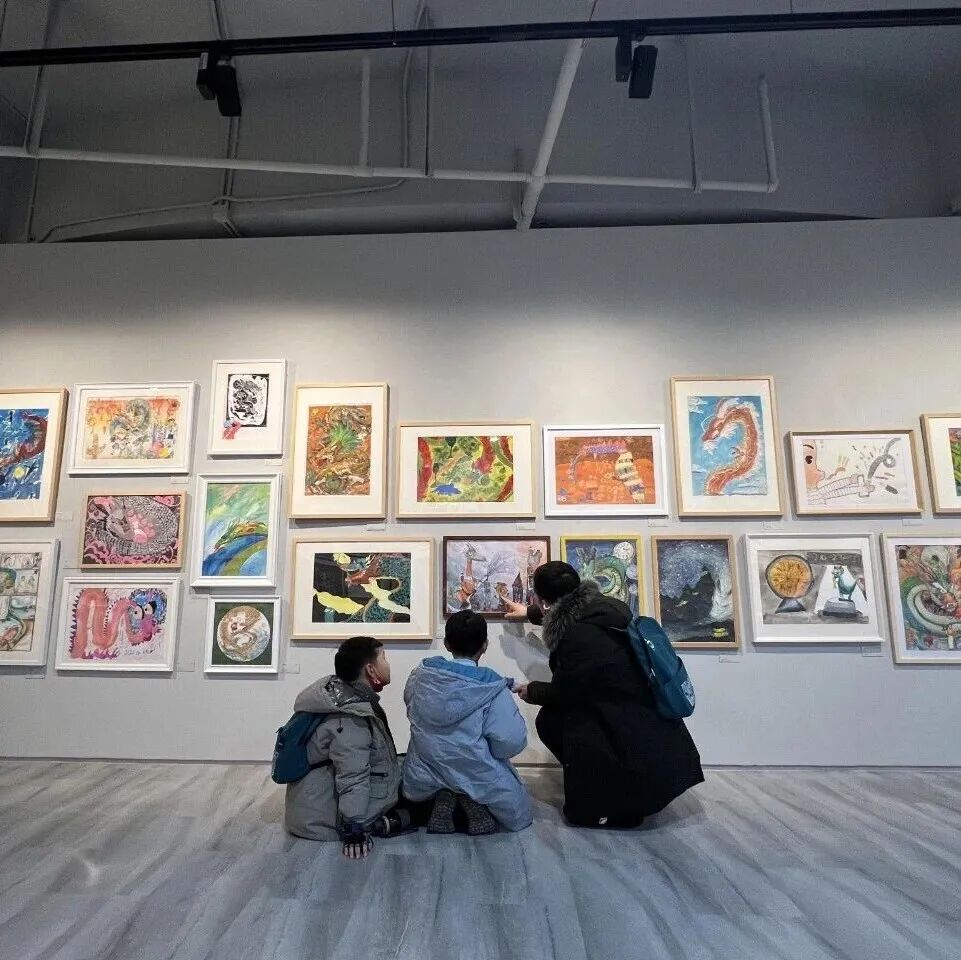 Three children crouch close to a wall filled with framed children's artworks, studying them together