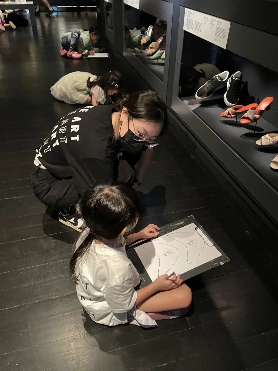 A Tree Art teacher guides a young girl sketching on a clipboard in a shoe exhibition gallery