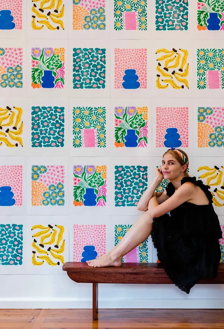 Liv Lee sitting on a wooden bench in front of a wall covered with her colorful banana, daisy, and vase illustrations