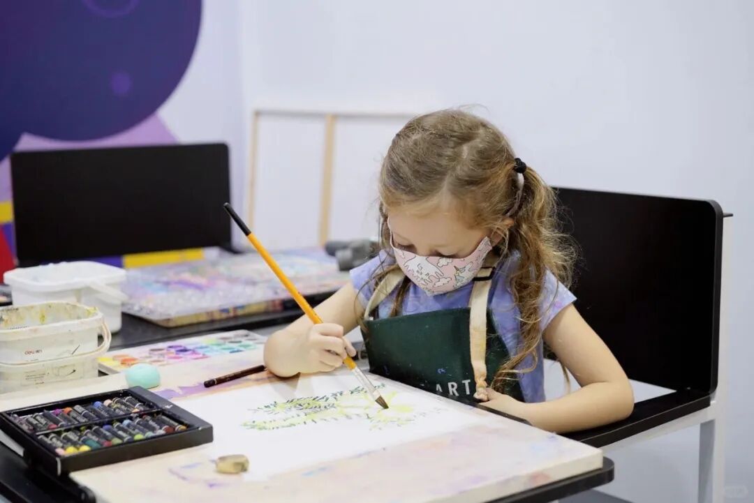 A young girl in a face mask paints with watercolors at a TreeArt studio table, paint palette and brushes in front of her